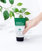 Hand holding a tube of Truecica Mineral 100 Calming Suncream with green leaves in the background